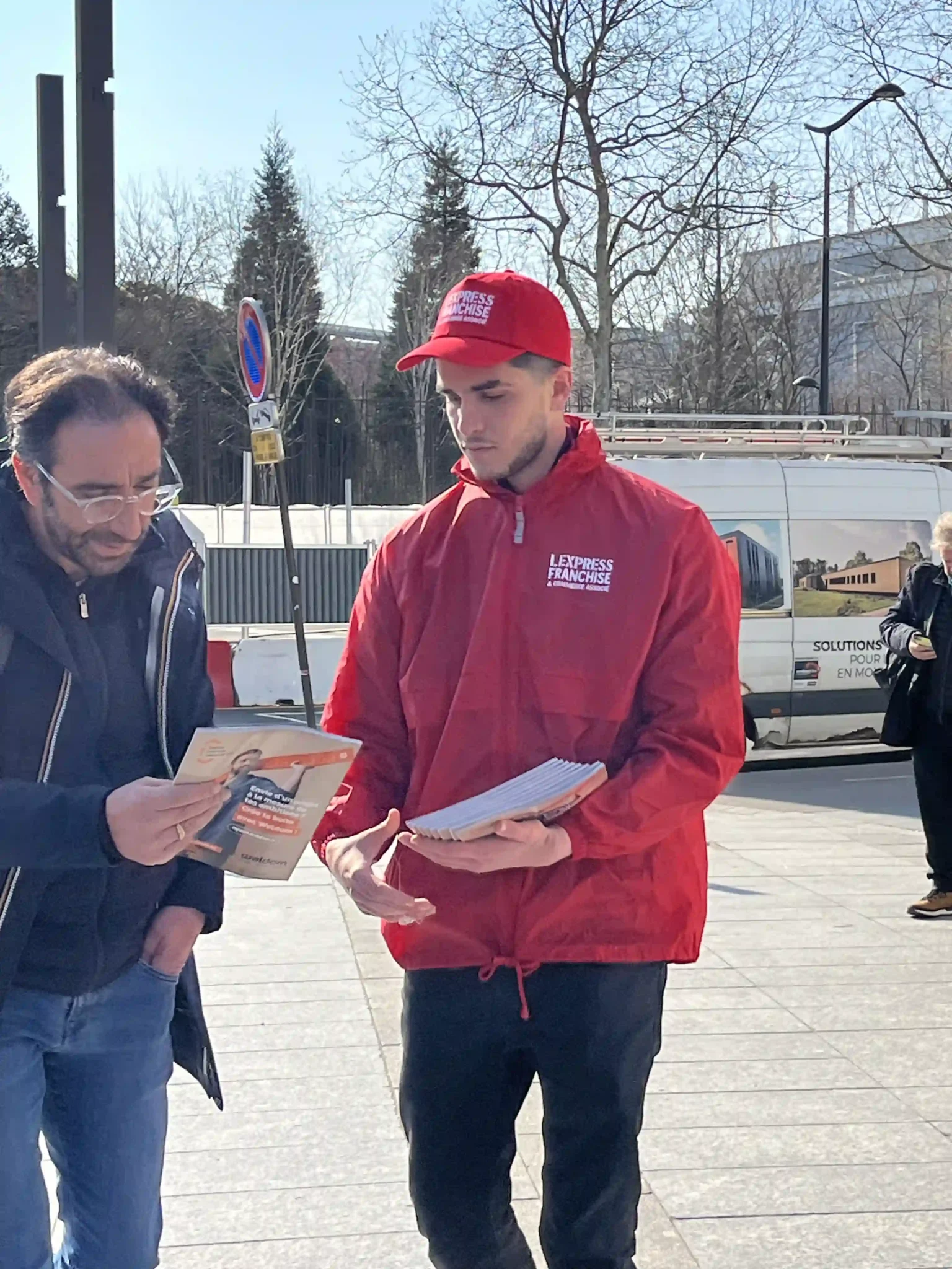 Un homme en tenue rouge, distribuant des flyers à un passant dans la rue lors d’une opération de street marketing.